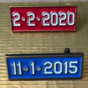 May include: Two enamel pins with gold trim. The top pin is red with white text that reads "2.2.2020". The bottom pin is blue with white text that reads "11.1.2015".
