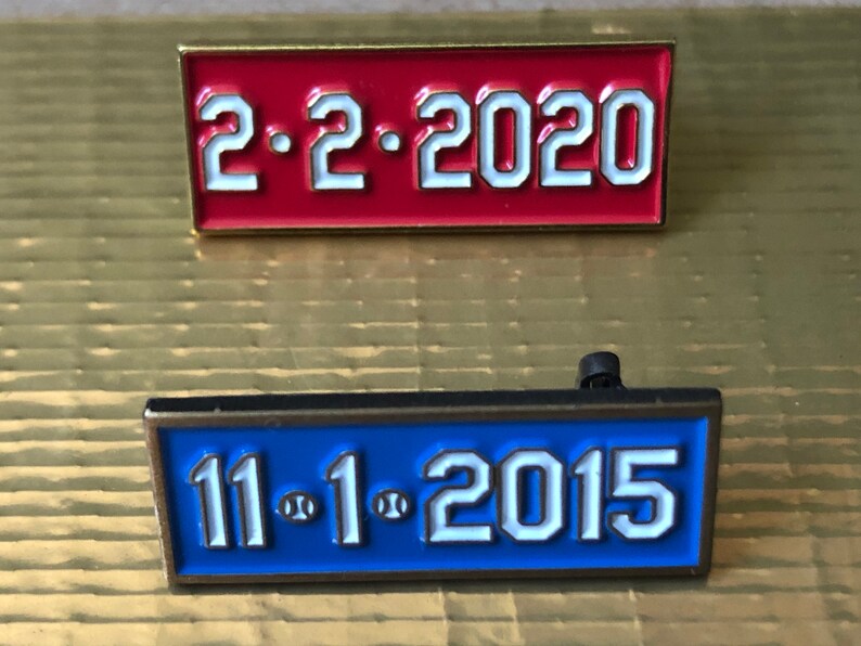 May include: Two enamel pins with gold trim. The top pin is red with white text that reads "2.2.2020". The bottom pin is blue with white text that reads "11.1.2015".