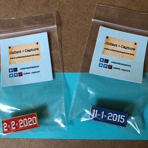 May include: Two small plastic bags containing enamel pins. The pins are shaped like license plates. The left pin is red and says "2-2-2020". The right pin is blue and says "11-1-2015".