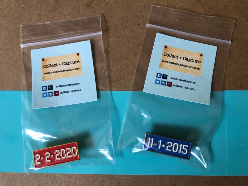 May include: Two small plastic bags containing enamel pins. The pins are shaped like license plates. The left pin is red and says "2-2-2020". The right pin is blue and says "11-1-2015".
