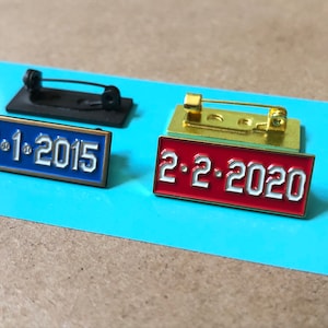 May include: Two enamel pins with gold-colored metal backing. The pin on the left is blue with white text that reads "11.1.2015". The pin on the right is red with white text that reads "2.2.2020".