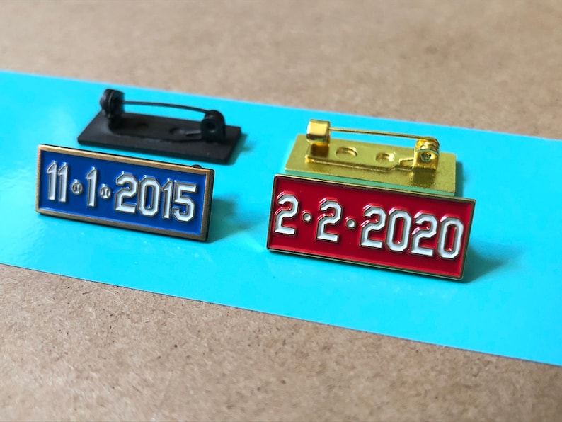 May include: Two enamel pins with gold-colored metal backing. The pin on the left is blue with white text that reads "11.1.2015". The pin on the right is red with white text that reads "2.2.2020".