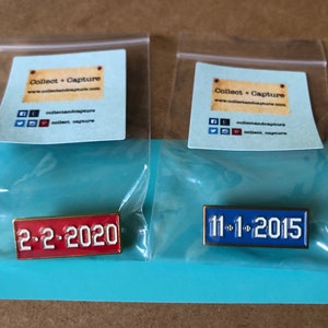 May include: Two enamel pins with gold trim. The left pin is red with white text that reads "2-2-2020". The right pin is blue with white text that reads "11-1-2015".
