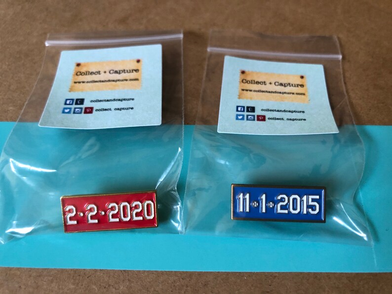 May include: Two enamel pins with gold trim. The left pin is red with white text that reads "2-2-2020". The right pin is blue with white text that reads "11-1-2015".