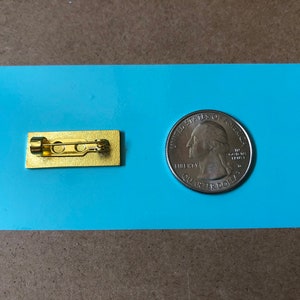 May include: A gold-colored metal pin back with a safety pin closure. The pin back is about the size of a US quarter.