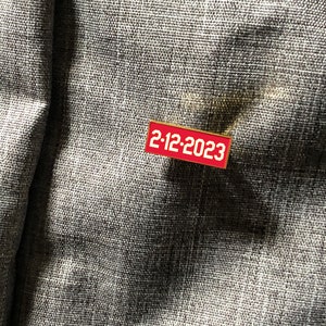 Op de afbeelding: Een rode emaille pin met een gouden rand en de tekst "2-12-2023" in wit.