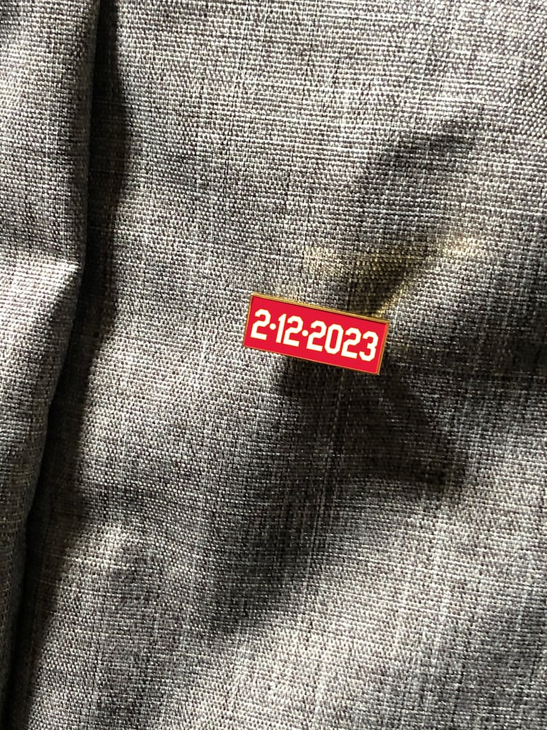 Op de afbeelding: Een rode emaille pin met een gouden rand en de tekst "2-12-2023" in wit.