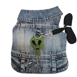 May include: A blue denim dog vest with an alien face and a UFO embroidered on the front. The vest has a black leash attached to it.