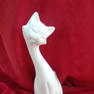 Könnte beinhalten: Eine weiße Keramik-Katzenfigur mit langem Hals und entspannter Pose. Die Katze hat geschlossene Augen und spitze Ohren. Die Figur steht vor einem roten Hintergrund.