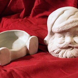 Puede incluir: Dos adornos navideños de cerámica sin pintar. Uno es una cabeza de Papá Noel con gorro y barba. El otro es un cuerpo de Papá Noel con piernas, diseñado para sostener un objeto pequeño. Ambos son blancos.