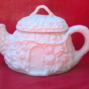 May include: White ceramic teapot shaped like a fairy house with a thatched roof and a small door. The teapot has a handle and a spout.