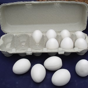 Könnte beinhalten: Eine Schachtel mit weißen Eiern, wobei einige Eier entnommen und auf eine blaue Oberfläche gelegt wurden. Die Schachtel besteht aus Pappe und hat eine hellgraue Farbe. Die Eier sind weiß und oval.