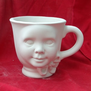 May include: White ceramic mug shaped like a child's face with a bow. The mug has a handle and is ready to be painted.