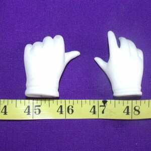 May include: Two white porcelain doll hands, one with a closed fist and the other with an open hand, are shown on a purple surface with a yellow measuring tape. The hands are approximately 4.5 inches long.
