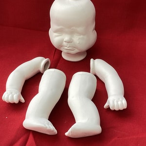 Puede incluir: Una cabeza de muñeca blanca sin terminar con una expresión de llanto y un cuerpo blanco con dos brazos y dos piernas. La muñeca está hecha de un material blanco liso y está lista para ser pintada.