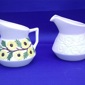 May include: Two white ceramic pitchers with different designs. One pitcher has a floral design with yellow flowers and green leaves. The other pitcher has a raised embossed design of flowers and leaves.