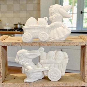 Könnte beinhalten: Zwei weiße Keramik-Osterhasenfiguren. Ein Hase zieht einen Wagen voller Eier, der andere schiebt einen Wagen mit Eiern. Die Figuren stehen auf einem Holzregal.