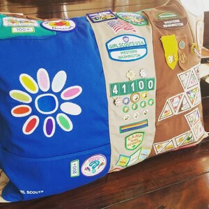 May include: A decorative pillow featuring a blue and tan design with Girl Scout patches and badges. The blue side has a large flower design and the words "Girl Scouts". The tan side has patches and pins.