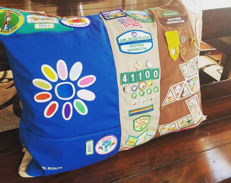 May include: A decorative pillow featuring a blue and tan design with Girl Scout patches and badges. The blue side has a large flower design and the words "Girl Scouts". The tan side has patches and pins.
