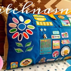 May include: Blue pillow with a large flower design and various patches. The pillow has a flower design with colorful petals and green leaves. Patches include numbers, badges, and text. The text "stitchnasnap" is written across the top.