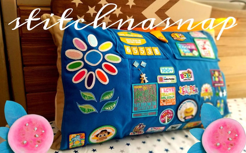 May include: Blue pillow with a large flower design and various patches. The pillow has a flower design with colorful petals and green leaves. Patches include numbers, badges, and text. The text "stitchnasnap" is written across the top.
