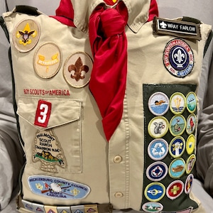 May include: A tan Boy Scout uniform pillow with a red neckerchief, a variety of merit badges, and the text "WRAY FARLOW" and "BOY SCOUTS OF AMERICA 1910-100 YEARS 2010".