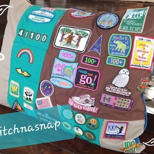May include: A custom pillow featuring Girl Scout patches. The pillow has a brown center with patches and a teal and blue border. Patches include text like "Girl Scouts USA", "Making Memories", and "U Go Girl!" The pillow is a unique keepsake.