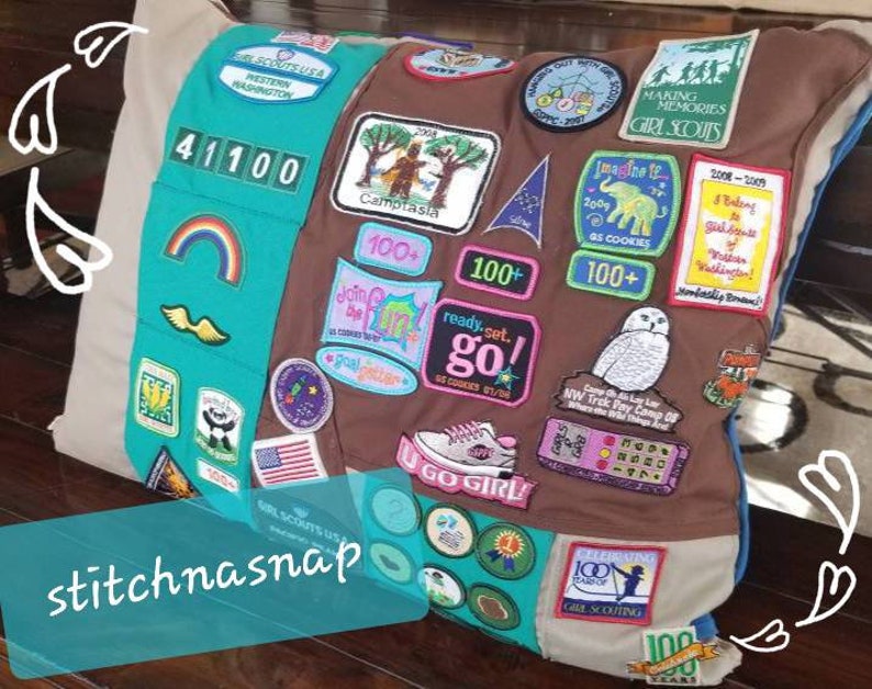 May include: A custom pillow featuring Girl Scout patches. The pillow has a brown center with patches and a teal and blue border. Patches include text like "Girl Scouts USA", "Making Memories", and "U Go Girl!" The pillow is a unique keepsake.
