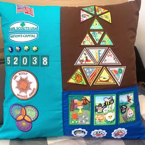 May include: A turquoise and brown pillow with Girl Scout patches and badges. The patches include a Girl Scouts USA Nation's Capital patch, a Girl Scout Bronze Award patch, and a variety of other patches with images of ladybugs, flowers, and other nature-themed designs. The pillow also has the numbers 52038 printed on it.