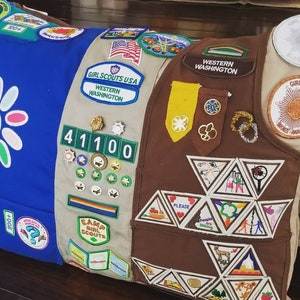 May include: A decorative pillow featuring a patchwork design with Girl Scout patches and badges. The pillow has sections of blue, tan, and brown fabric, adorned with various embroidered patches, including the words "Girl Scouts USA" and "Western Washington."
