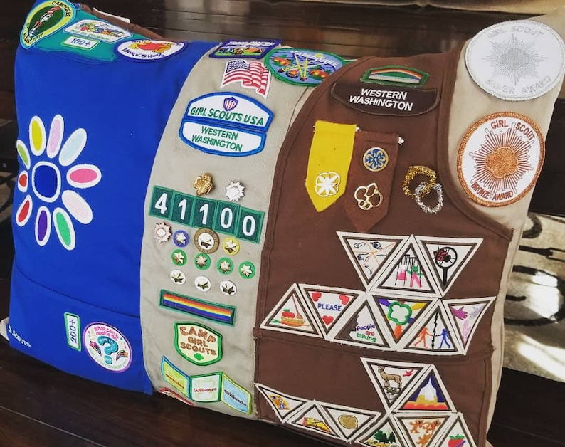 May include: A decorative pillow featuring a patchwork design with Girl Scout patches and badges. The pillow has sections of blue, tan, and brown fabric, adorned with various embroidered patches, including the words "Girl Scouts USA" and "Western Washington."