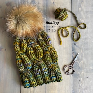 May include: A green, yellow and purple knitted hat with a brown faux fur pom pom. The hat is lying on a wooden surface with yarn and scissors.