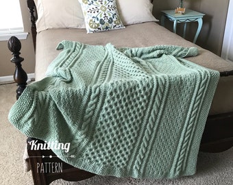 May include: A sage green knitted blanket featuring a textured pattern of vertical cable stitches and honeycomb-like sections. The blanket is draped over a bed and includes the text Knitting PATTERN printed in white on the lower left corner.