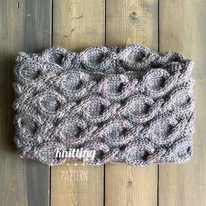 Può includere: Un primo piano di un collo a maglia con un motivo a treccia in tonalità di grigio e viola. Il collo è disteso su una superficie di legno. Il testo "Knitting Pattern" è visibile nella parte inferiore dell'immagine.