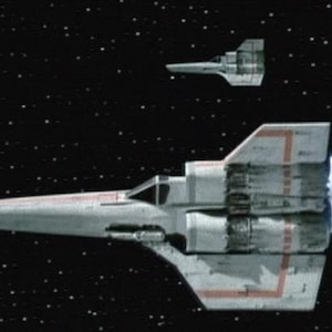 May include: Two white spacecraft with red detailing are shown against a starry black backdrop. The larger ship is in the foreground, with a smaller ship above it. The larger ship has a long, pointed nose and a rectangular body.