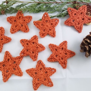 Könnte beinhalten: Acht gehäkelte orangefarbene Stern-Ornamente auf weißem Hintergrund. Die Sterne sind aus einem strukturierten Garn gefertigt und haben eine fünfzackige Sternform.