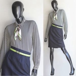 May include: A black and white striped long-sleeved blouse with a green and white patterned scarf tied around the collar. The blouse has a green belt with a silver buckle. The model is wearing a dark blue skirt.