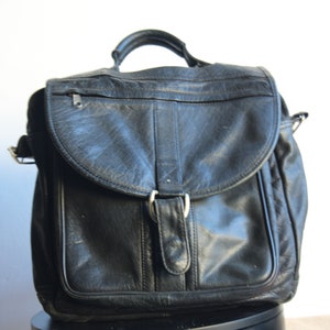 以下が含まれることがあります： 大きなフラップとバックル留めがついた黒い革のメッセンジャーバッグ。バッグにはショルダーストラップとトップハンドルが付いています。