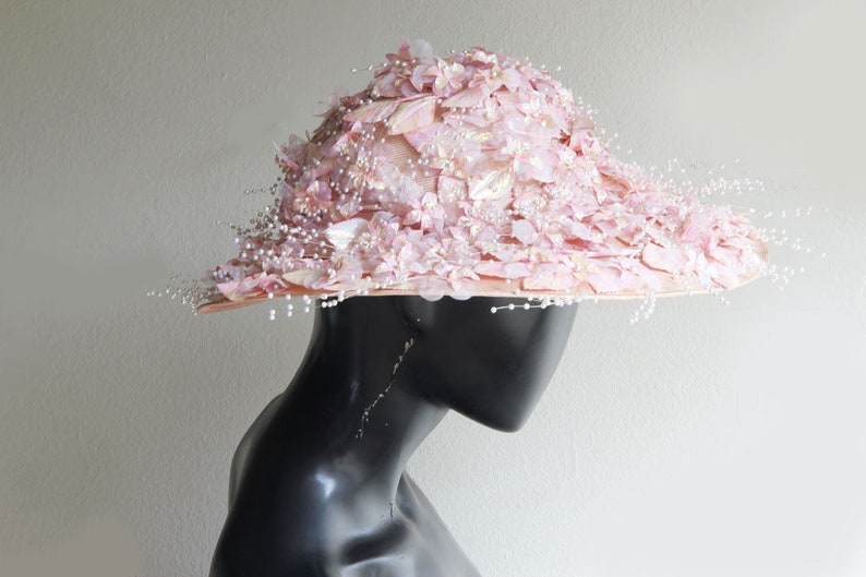 Puede incluir: Un sombrero de ala ancha con un dise&ntilde;o floral rosa y blanco. El sombrero est&aacute; decorado con cuentas de imitaci&oacute;n de perlas y tiene una est&eacute;tica delicada y femenina.