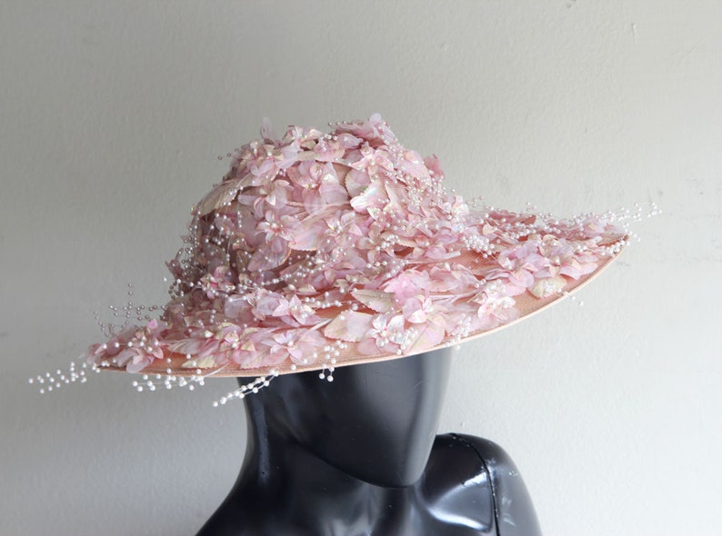 Puede incluir: Un sombrero de ala ancha con una base rosa y una corona cubierta de delicadas flores rosas y cuentas de perlas.