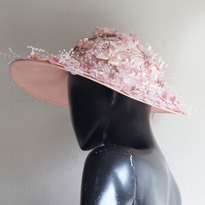 Puede incluir: Un sombrero rosa de ala ancha con un gran dise&ntilde;o floral decorativo. El sombrero est&aacute; adornado con delicadas cuentas blancas y plumas.