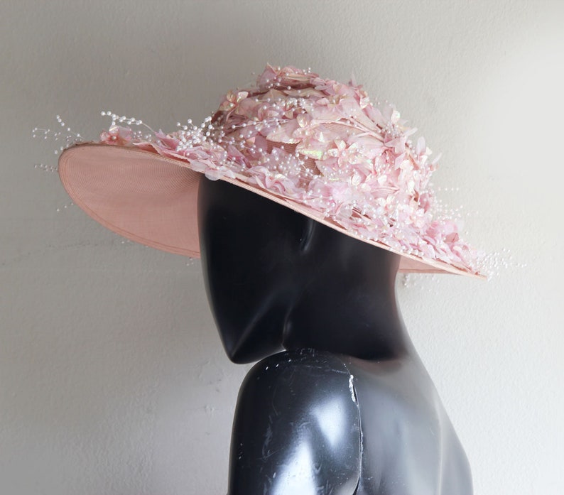 Puede incluir: Un sombrero rosa de ala ancha con un gran dise&ntilde;o floral decorativo. El sombrero est&aacute; adornado con delicadas cuentas blancas y plumas.