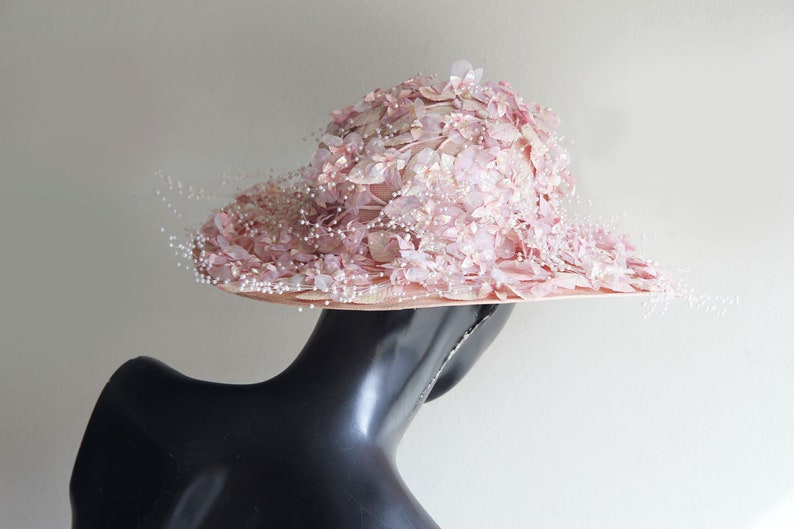 Puede incluir: Un sombrero rosa con un ala ancha y un dise&ntilde;o floral. El sombrero est&aacute; decorado con peque&ntilde;as cuentas blancas y tiene un velo delicado.