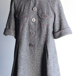 Puede incluir: Una chaqueta gris con capucha, costuras rosas y varios botones. La chaqueta tiene una forma ligeramente acampanada y mangas cortas.
