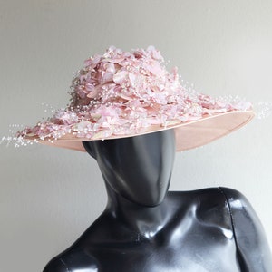 Puede incluir: Un sombrero de ala ancha con una base rosa y una gran flor rosa como adorno. El sombrero est&aacute; decorado con peque&ntilde;as cuentas blancas y tiene un dise&ntilde;o delicado y femenino.