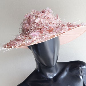 Puede incluir: Un sombrero rosa de ala ancha con un dise&ntilde;o floral y detalles de perlas. El sombrero est&aacute; hecho de un material suave y delicado y tiene una ala ancha que se puede peinar de varias maneras.