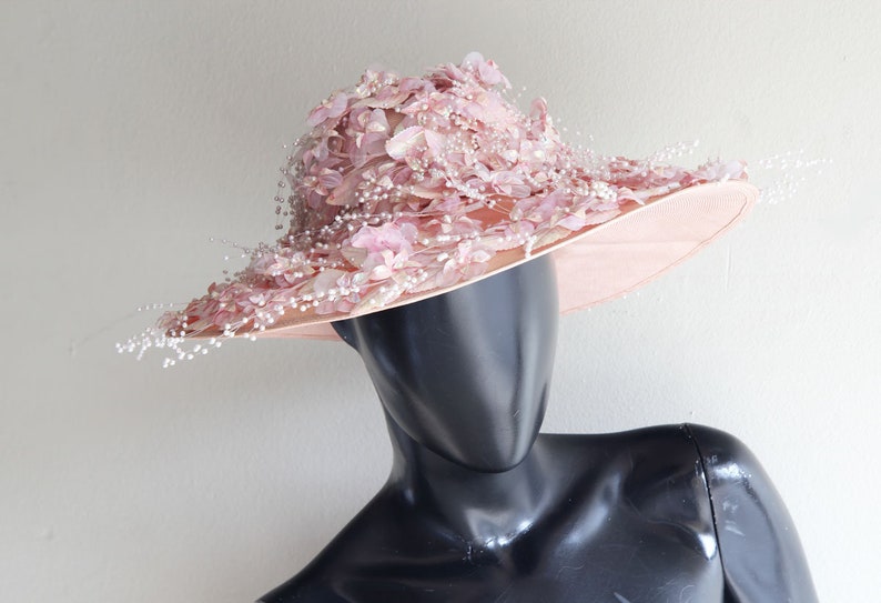 Puede incluir: Un sombrero rosa de ala ancha con un dise&ntilde;o floral y detalles de perlas. El sombrero est&aacute; hecho de un material suave y delicado y tiene una ala ancha que se puede peinar de varias maneras.