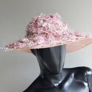 Puede incluir: Un sombrero de ala ancha de color rosa con un gran dise&ntilde;o floral y detalles de perlas.