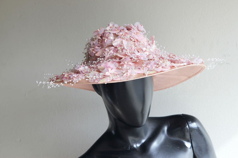 Puede incluir: Un sombrero de ala ancha de color rosa con un gran dise&ntilde;o floral y detalles de perlas.