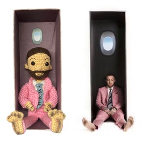 May include: A crocheted doll wearing a pink suit sits inside a box with a porthole. The doll has brown hair, a beard, and a blue tie. The box is brown with a purple interior.